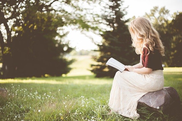 žena s dlouhými blonďatými vlasy sedí na kameni v přírodě, čte si knihu